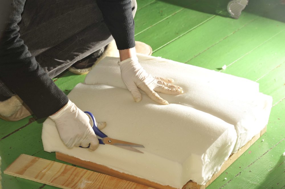A woman reupholsters a kitchen stool using scissors to trim the foam